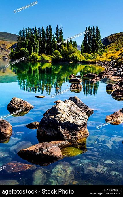 Cypress alley and huge basalt boulders reflected in the lake. The surroundings of the Cromwell. Magic New Zealand. The concept of ecological