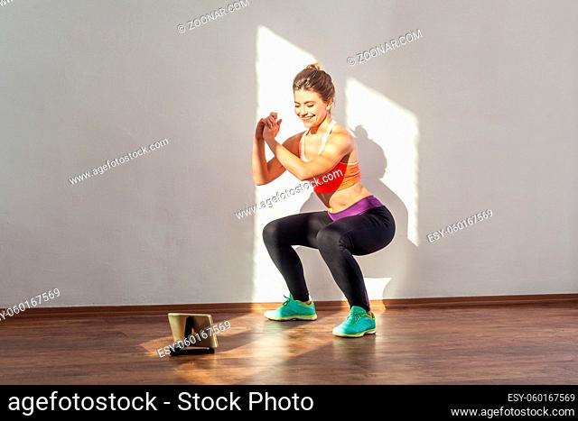 Positive sportive woman with bun hairstyle and in tight sportswear doing squatting sit-up exercise while watching training video on tablet