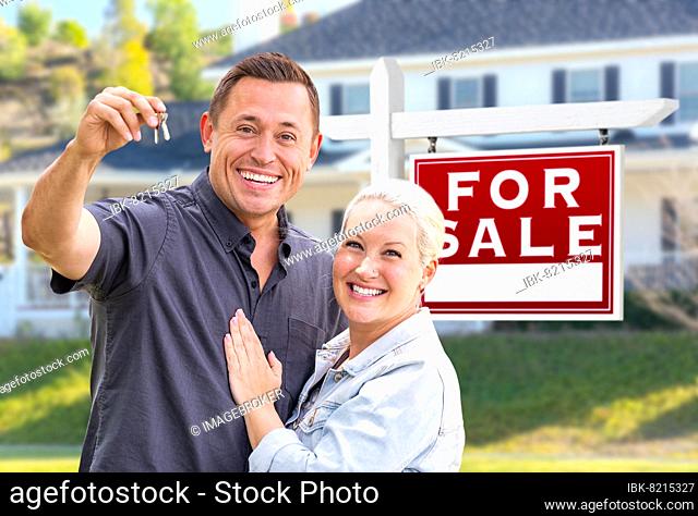 Young adult couple with house keys in front of home and for sale real estate sign
