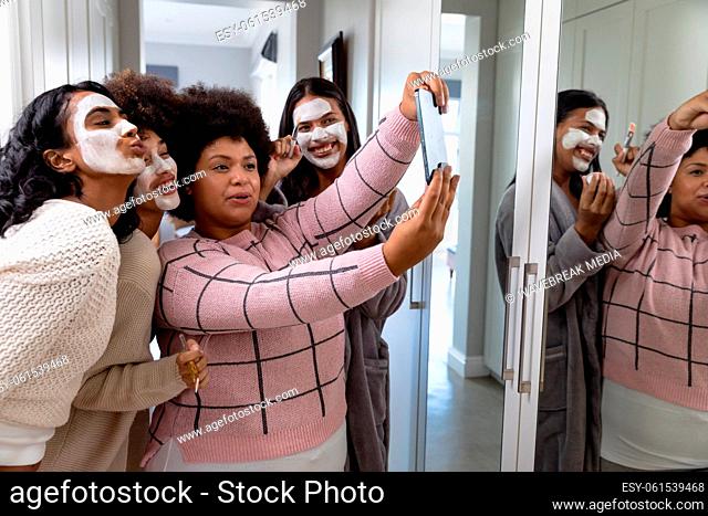 Happy biracial woman taking selfie over cellphone with female friends applied beauty cream on faces