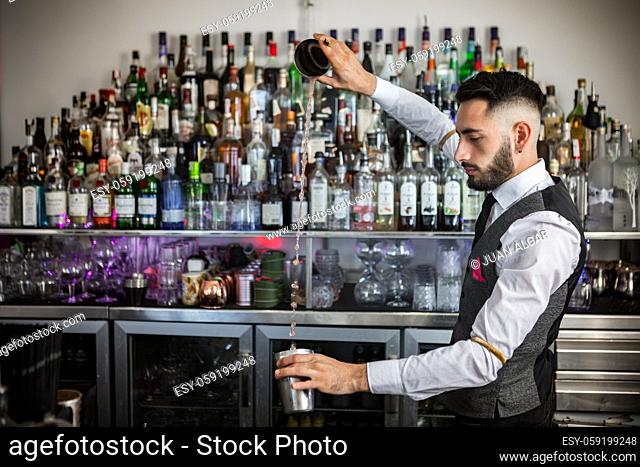 Side view of young bearded male barkeeper in elegant uniform pouring alcohol drink from shaker into stainless cup while preparing order at bar counter