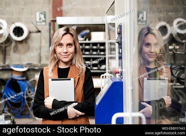 Smiling businesswoman with tablet PC standing by machine at industry