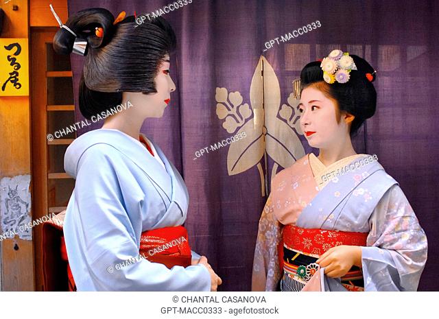 PORTRAIT OF A GEIKO GEISHA, ON THE LEFT, AND A MAIKO APPRENTICE WEARING A KIMONO OBEBE HELD CLOSED BY A WIDE BELT OBI, IN FRONT OF A GEISHAS’ HOUSE OKIYA
