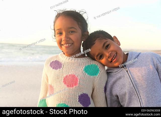 Portrait of smiling biracial girl with brother leaning head on her shoulder at beach during sunset