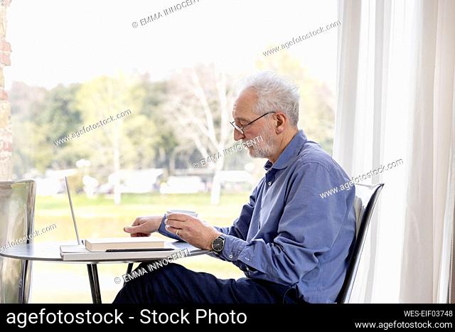 Senior man with laptop sitting at table in hotel apartment