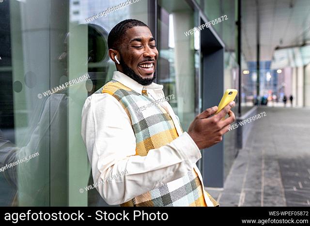 Happy man surfing net through smart phone in front of glass wall