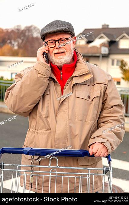Senior with winter cloth using cell phone while he is pushing is shopping cart on parking area