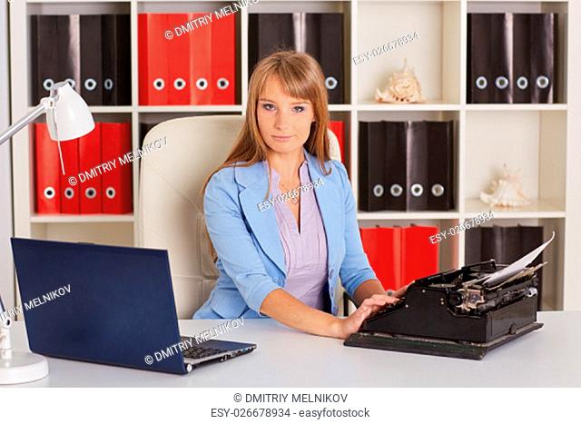 Young business woman with notebook and old typewriter sits on workplace in the office. Progressing of technology
