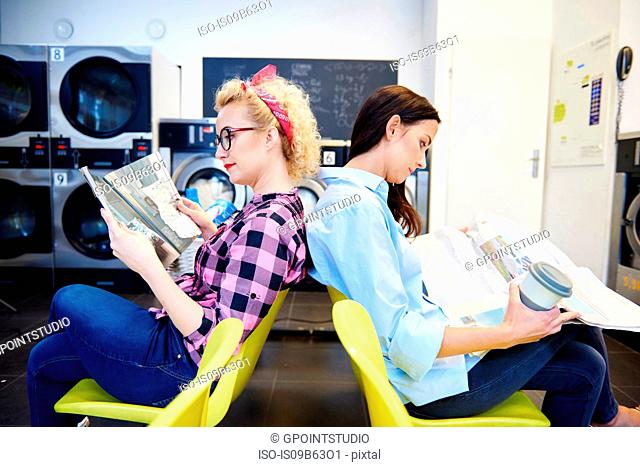 Two women reading newspapers back to back in laundrette