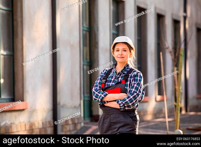 Portrait of happy, self-confident female worker in white hard hat and working uniform, posing with hands folded on chest