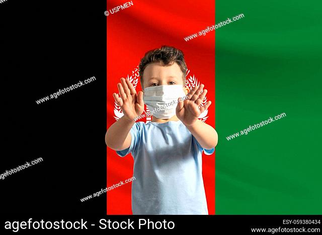 Little white boy in a protective mask on the background of the flag of Afghanistan. Makes a stop sign with his hands, stay at home Afghanistan