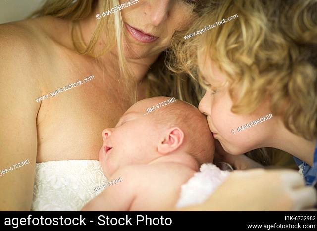 Beautiful young mother holds newborn baby girl as brother kisses her head
