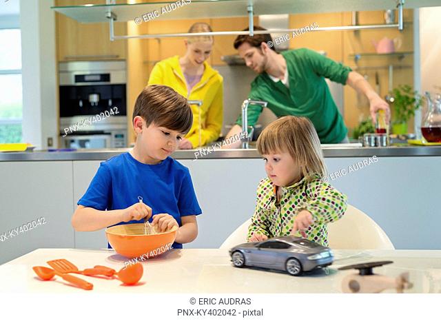 Little girl with a toy car looking at his brother cooking in the kitchen