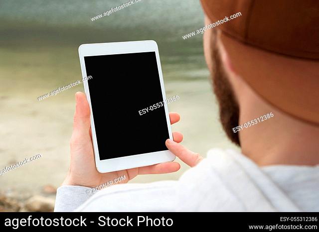 Close-up of a horde in a brown cap in the open air holds a white tablet pc in his hands. A bearded man looks at the tablet