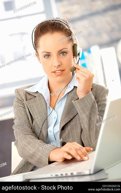 Attractive customer servicer working in bright office, using headphones and laptop