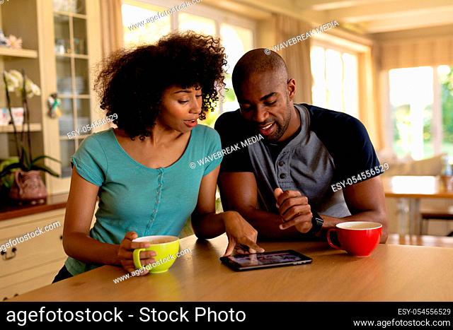Front view of a mixed race couple sitting at the counter in the kitchen with cups of coffee, smiling and using a tablet computer