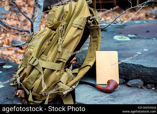 Close up advertising style shot of a green army tactical backpack, a paper notebook and a vintage pipe disposed on rock steps in the woodlands