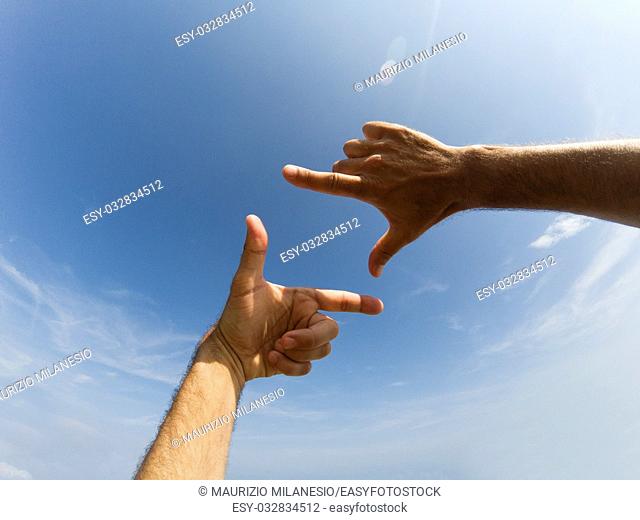 Two hands form with a fingers a frame in front of a blue sky with clouds and reflection