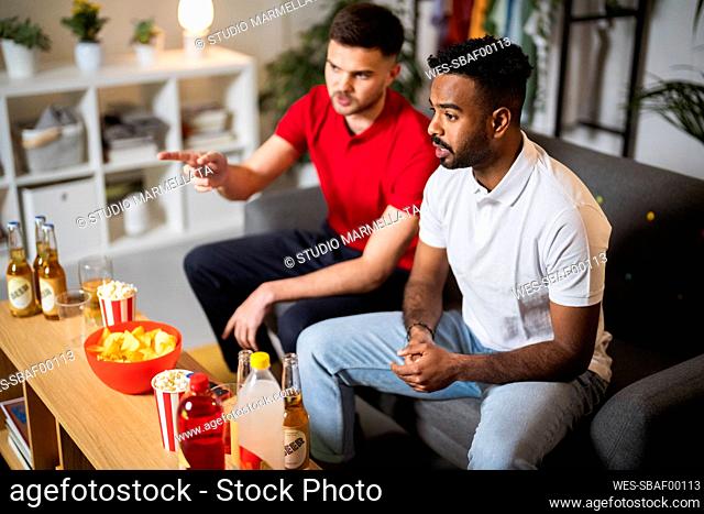 Young man talking with friend watching football match sitting on sofa at home