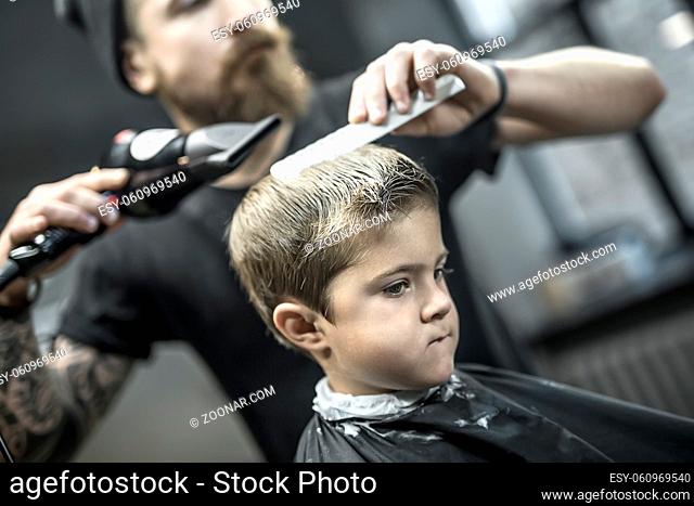 Handsome kid in a black salon cape in the barbershop. Bearded barber with a tattoo in a black T-shirt does kid the hairstyle with the help of the comb and the...