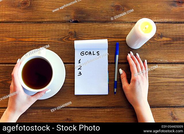 Female hand with pen writing goals in empty notebook on wooden table background with coffee and burning candle