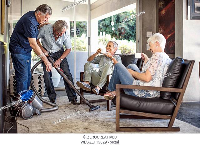 Two senior men vacuming house together while their wifes watching