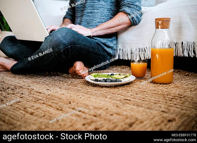 Young man working on laptop while sitting by juice and fruit plate on floor at home