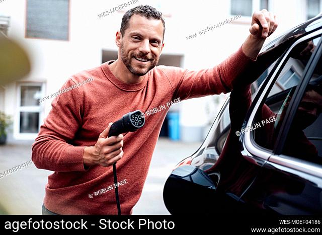 Smiling man holding charging cable leaning on electric car