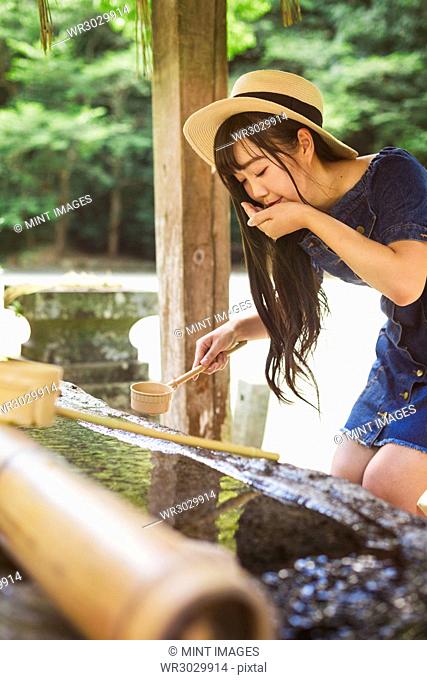 Young woman wearing blue dress and hat using bamboo water hand washing basins at Shinto Sakurai Shrine, Fukuoka, Japan