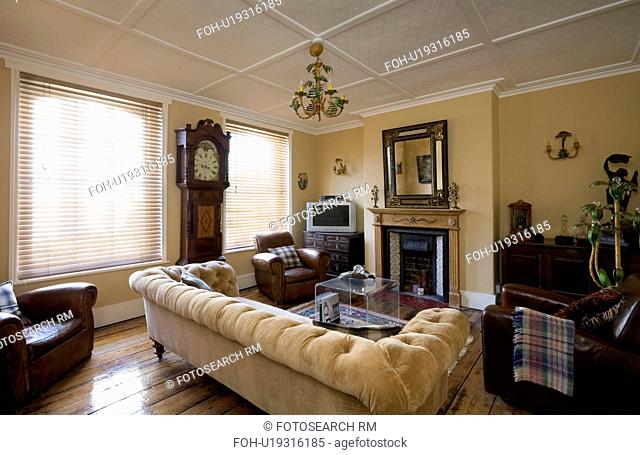 Neutral velour chesterfield sofa in traditional living room with longcase clock between windows with slatted blinds