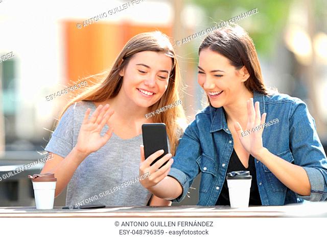 Two happy friends greeting having a video call on smart phone sitting in a park