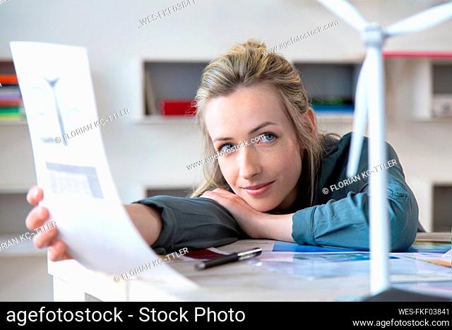 Portrait of woman leaning on desk in office with paper and wind turbine model