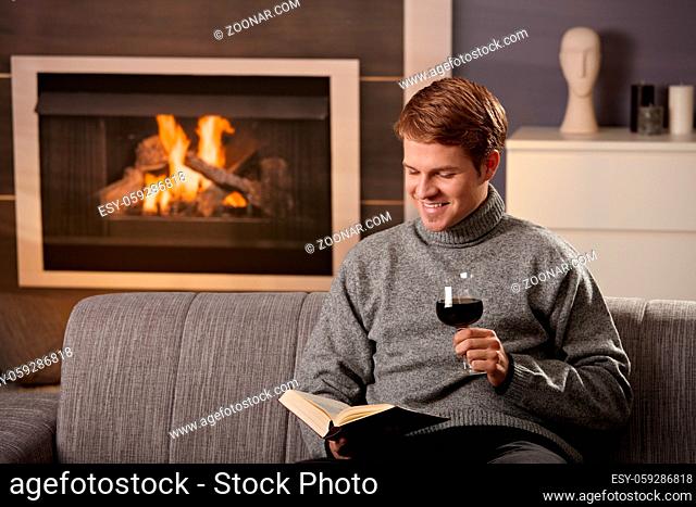 Young man sitting on sofa at home on a cold winter day, reading book in front of fireplace, tasting red wine