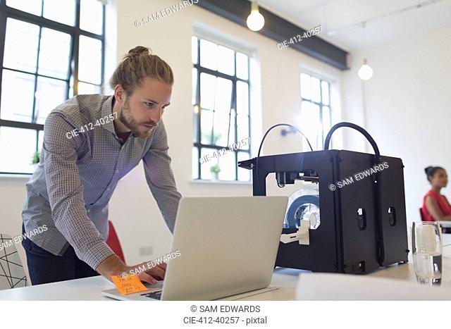 Male designer working at laptop next to 3D printer in office