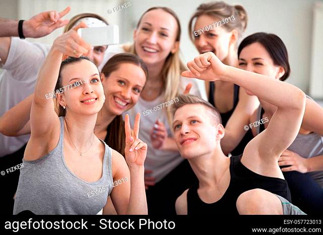 Group millennial positive laughing sportive athletic girls and guys at gym training studio. People smiling make peace sign symbol taking selfie photo feels...
