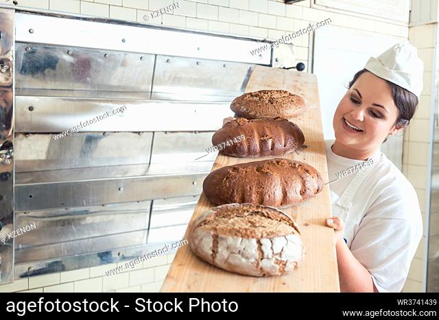 Baker woman presenting bread on board in bakery looking proudly into the camera