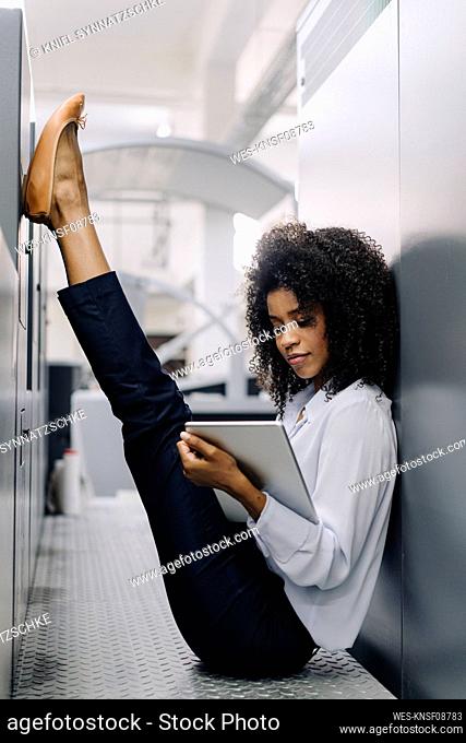 Businesswoman working on digital tablet while sitting with feet up amidst machines at industry