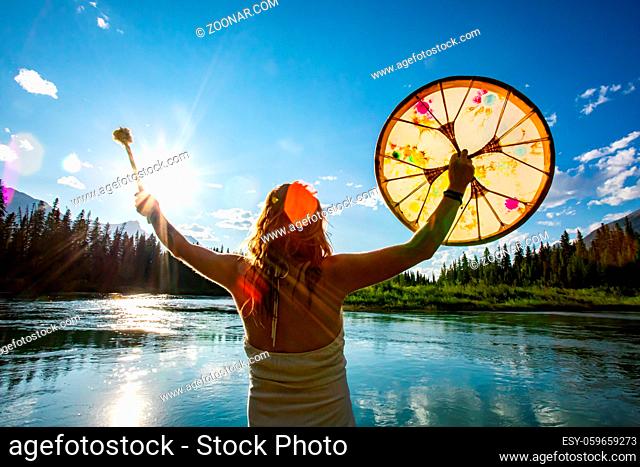 A creative lens flare is seen over a backlit woman holding a native drum and beater up, celebrating traditional culture and natural surroundings