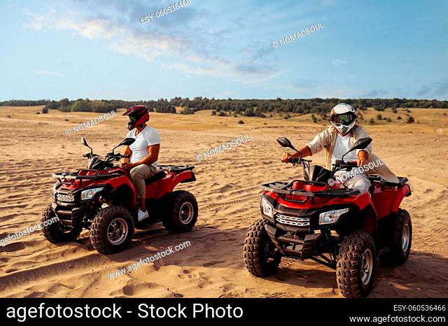 Two men in helmets ride on atv in desert. Male persons on quad bikes, sandy race, dune safari in hot sunny day, 4x4 extreme adventure, quad-biking