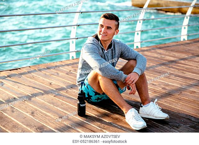 Young stylish sportsman listening to music in earphones, resting on pier after workout, with bottle of water. Dressed in sportswear and sneakers