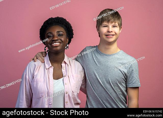 Friendship. Smiling embracing dark-skinned woman and guy with down syndrome looking at camera standing on light background