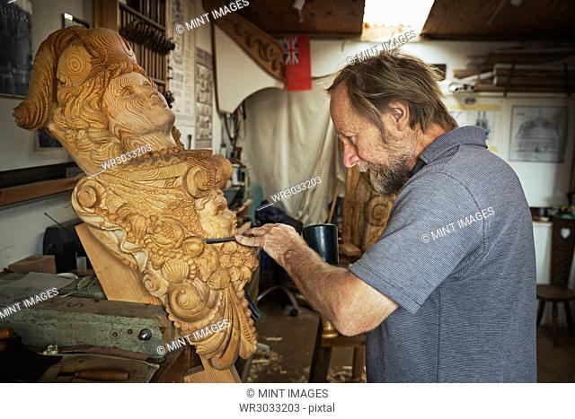 A craftsman, a wood carver working on a wooden female ship's figurehead held in a vice on the workbench of his workshop