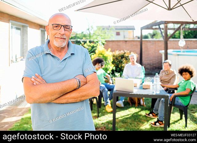 Senior man standing with arms crossed while family in background at backyard