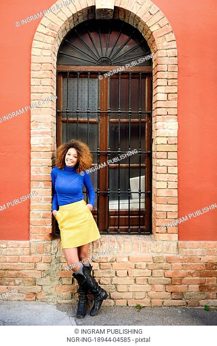 Beautiful young African American woman, model of fashion, smiling with afro hairstyle and green eyes wearing blue sweater and yellow skirt in urban background