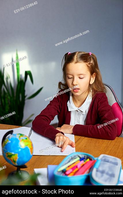 Schoolgirl sit at desk doing homework reading, homeschooling. Small junior girl engaged in distant learning get remote elementary education and preparing for...