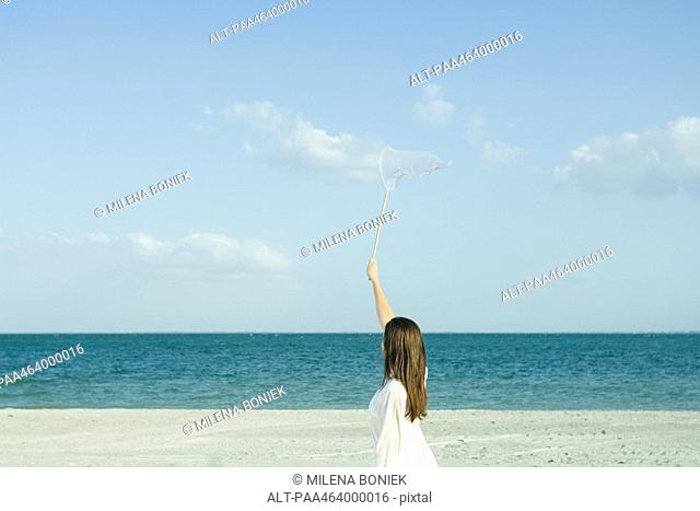 Woman on beach catching clouds with butterfly net, rear view, optical illusion