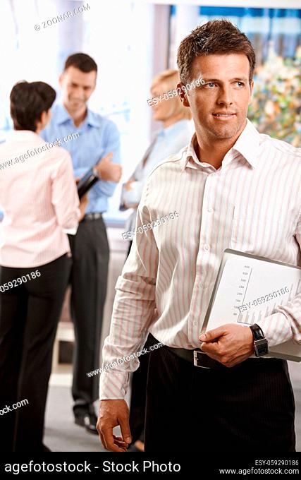 Portrait of businessman in office, holding notepad. Colleagues talking in the background
