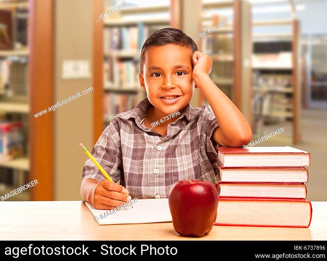 Adorable hispanic boy with books, apple, pencil and paper studying at the library