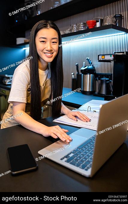 Smiling cheerful cute young cafe employee touching the account book while working on the laptop