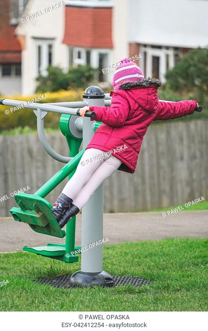 Little Caucasian girl exercising on the outdoor training machine on a chilly autumn day